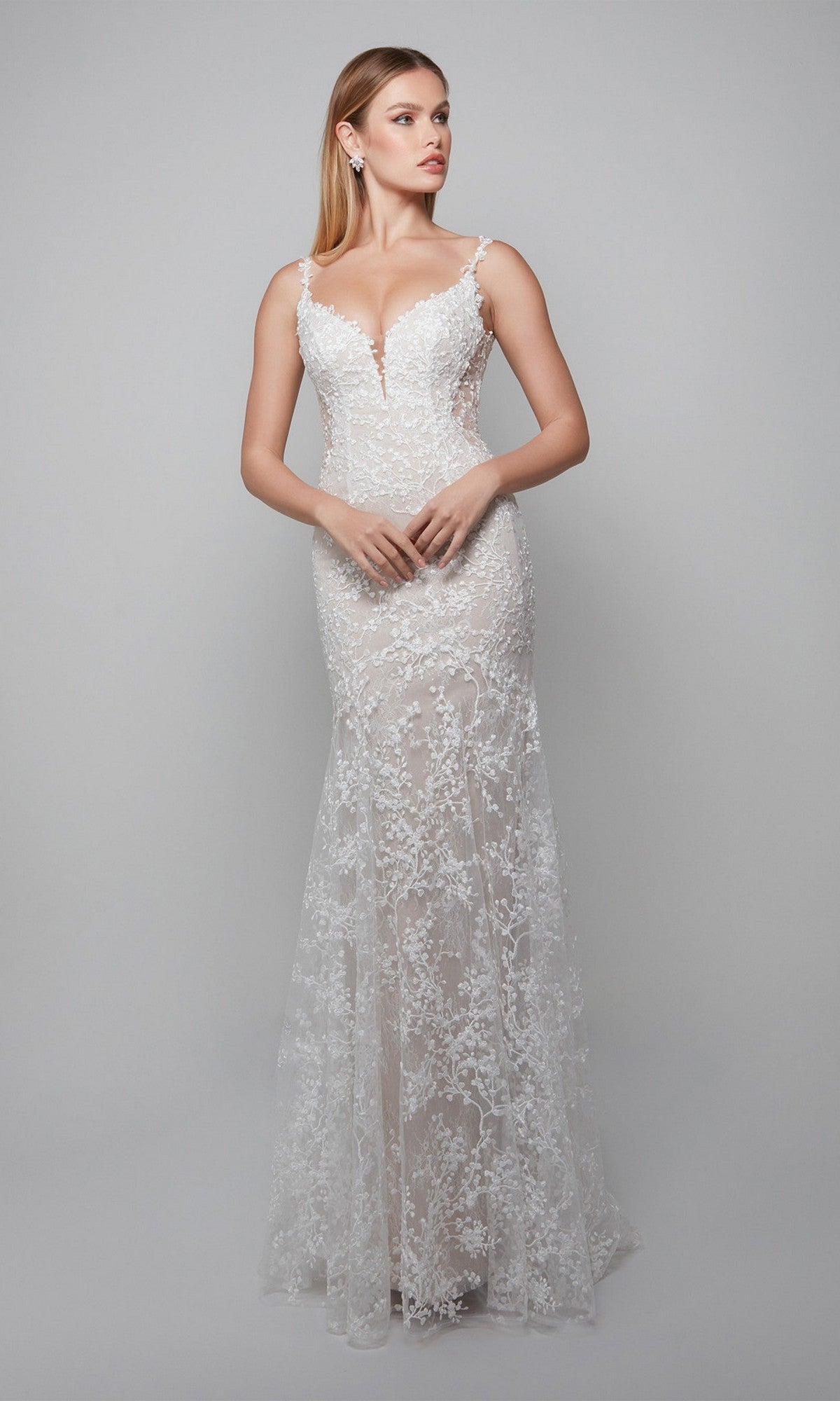 A woman wears the Alyce 7077 Long White Dress, an ivory formal gown with a sleeveless lace bodice, deep neckline, and floor-length skirt, standing against a plain gray background.